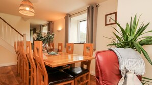 A dining area with a wooden table, chairs, a vase with flowers, a plant, and a window with curtains.