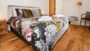 Uma cama bem arrumada com roupa de cama floral, uma mesa de cabeceira de madeira, uma cadeira e um abajur.
