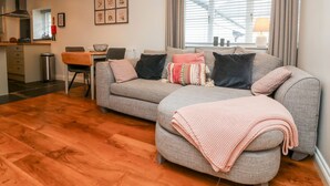 Uma sala de estar moderna com um sofá cinzento, uma área de jantar e uma cozinha ao fundo.