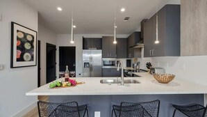 A modern kitchen with a central island, stainless steel appliances, pendant lighting, and black wire mesh chairs. - Tune - Odyssey - Pool & Gym - Mins to Broadway! (Nashville)