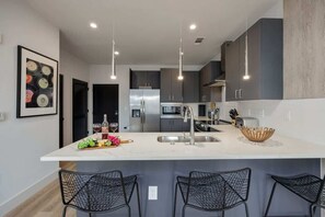 A modern kitchen with a central island, stainless steel appliances, pendant lighting, and black wire mesh chairs.