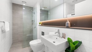 Un baño moderno con cabina de ducha de vidrio, inodoro blanco, lavamanos con espejo y una planta en maceta.