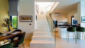 Une cuisine moderne avec des meubles blancs, un escalier en verre et un coin repas doté de chaises recouvertes d’un tissu vert.