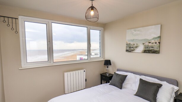 A bedroom with a large bed, a radiator, a nightstand with a lamp, a wall-mounted picture, and a window with a view of the beach. - Apt 10 @ Hunter's Quay (Bridlington)