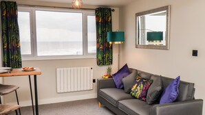 A living room with a grey sofa, a small table, a chair, a floor lamp, a radiator, and a window with curtains. - Apt 10 @ Hunter's Quay (Bridlington)
