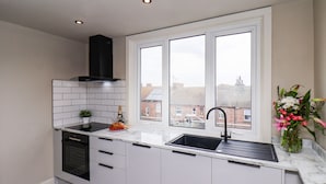 A modern kitchen with white cabinets, a black range hood, a built-in oven, a sink, and a window with a view of buildings. - Apt 10 @ Hunter's Quay (Bridlington)