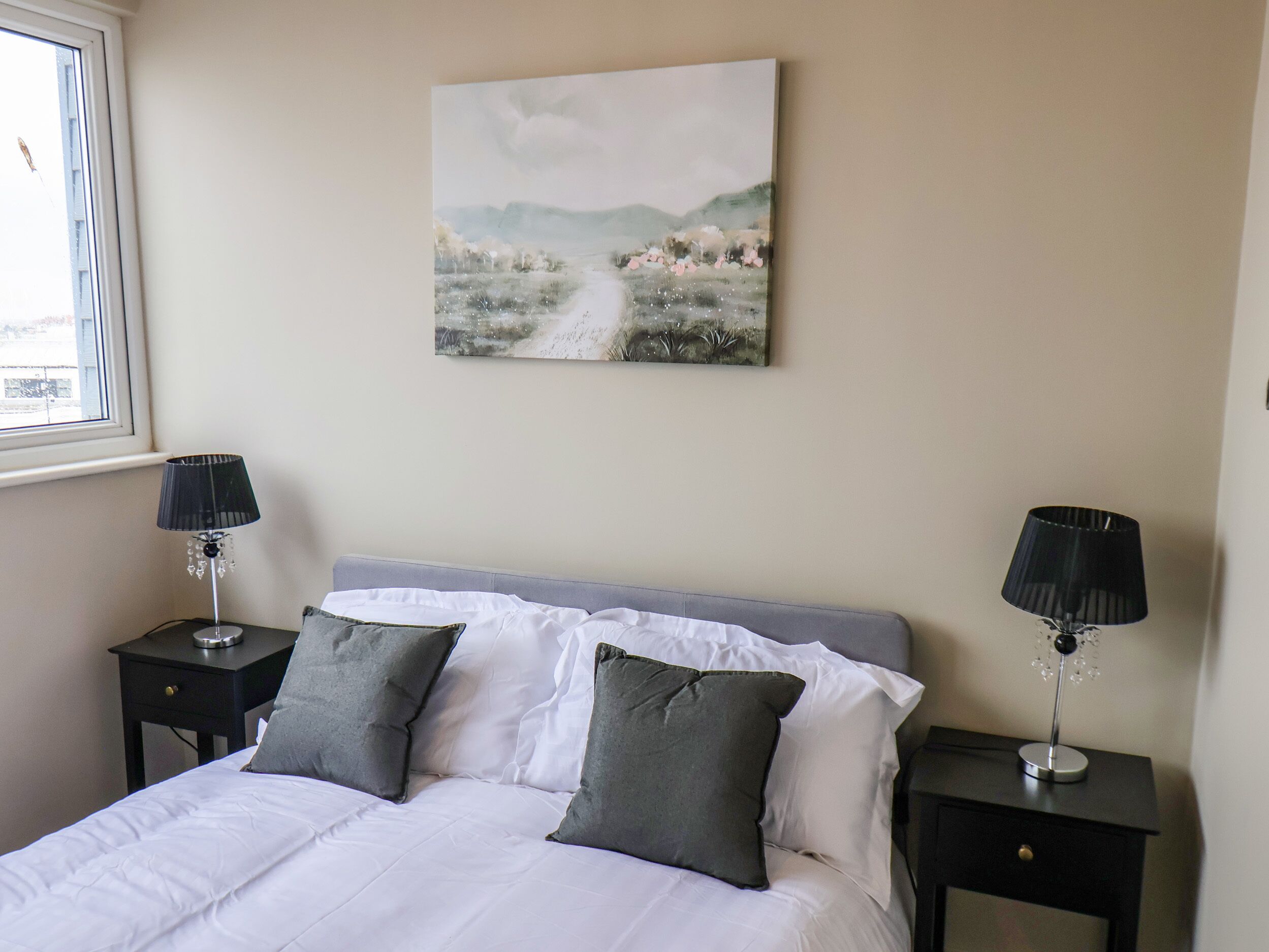 A bedroom with a bed, two bedside tables, a window, and a painting on the wall.