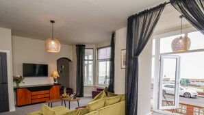 Ein modernes Wohnzimmer mit einem grünen Sofa, einem Holzesstisch mit Frühstücksgebäck, einem Fernseher und einem großen Fenster mit Blick auf einen Parkplatz.