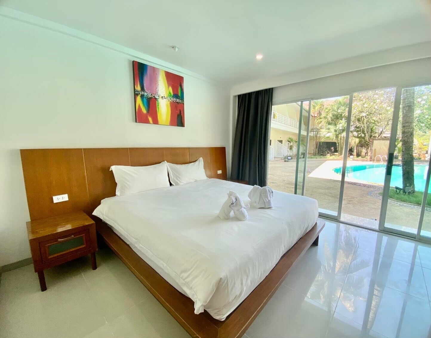 A large bed with white linens, a wooden headboard, and a nightstand with a drawer. A colorful abstract painting is on the wall.