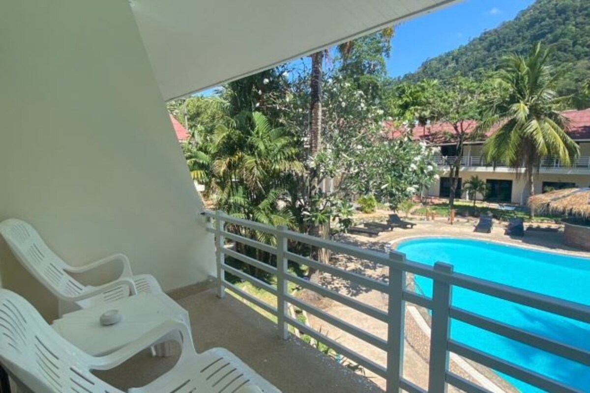 A balcony with white plastic chairs and a view of a swimming pool surrounded by palm trees and a mountain backdrop.