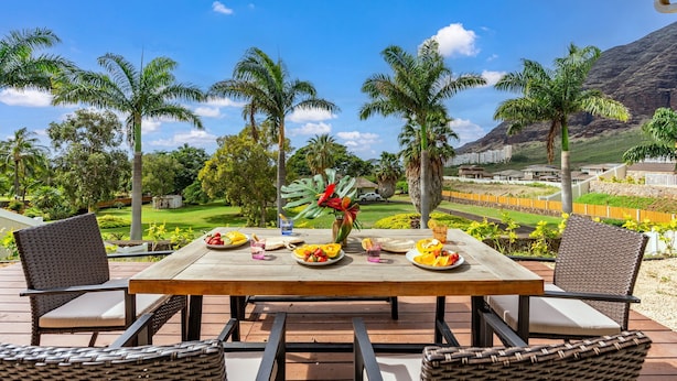 A wooden deck with a table set for a meal, surrounded by wicker chairs, overlooking a lush landscape with palm trees and a distant mountain. - Amazing Ocean Views, Game Room & Modern Design | Aloha Breeze 932 by AvantStay (Waianae)