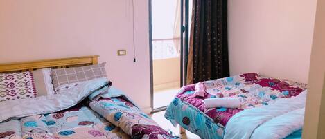 Un dormitorio con dos camas, cada una con ropa de cama estampada. Hay una ventana con cortinas y una puerta que da al exterior.