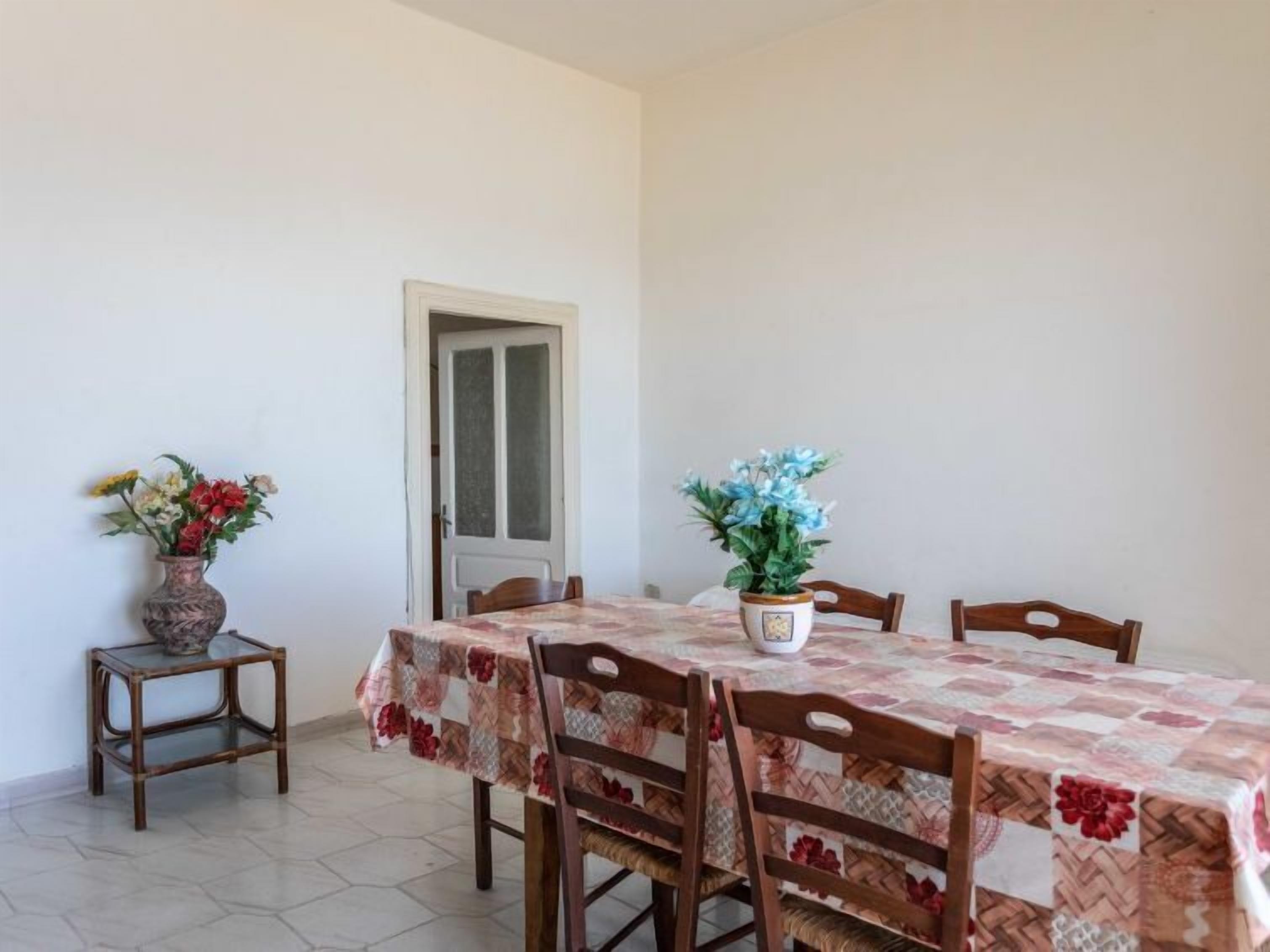 A dining area with a table covered in a floral tablecloth, surrounded by wooden chairs. There are two potted plants on the table, one with yellow and white flowers and the other with blue flowers. A door is visible in the background.