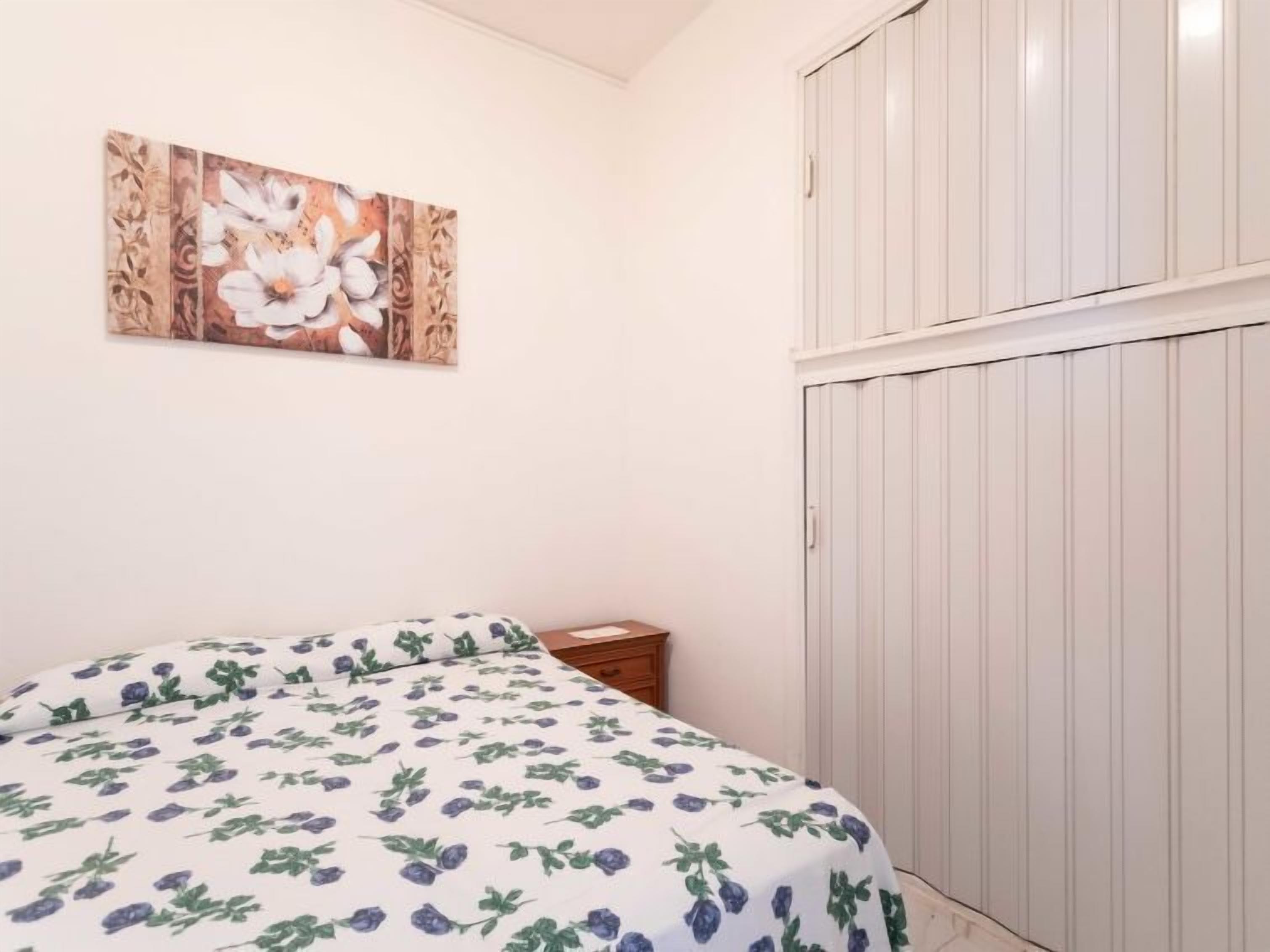 A bedroom with a bed covered in a floral-patterned duvet, a wooden nightstand, a white wardrobe, and a floral artwork on the wall.