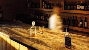 A bar area with wooden shelves stocked with liquor bottles, a bar counter with a bottle of Jack Daniel's whiskey, and pendant lighting.