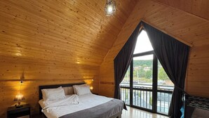 A wooden cabin bedroom with a large bed, a nightstand, a lamp, and a window with curtains.