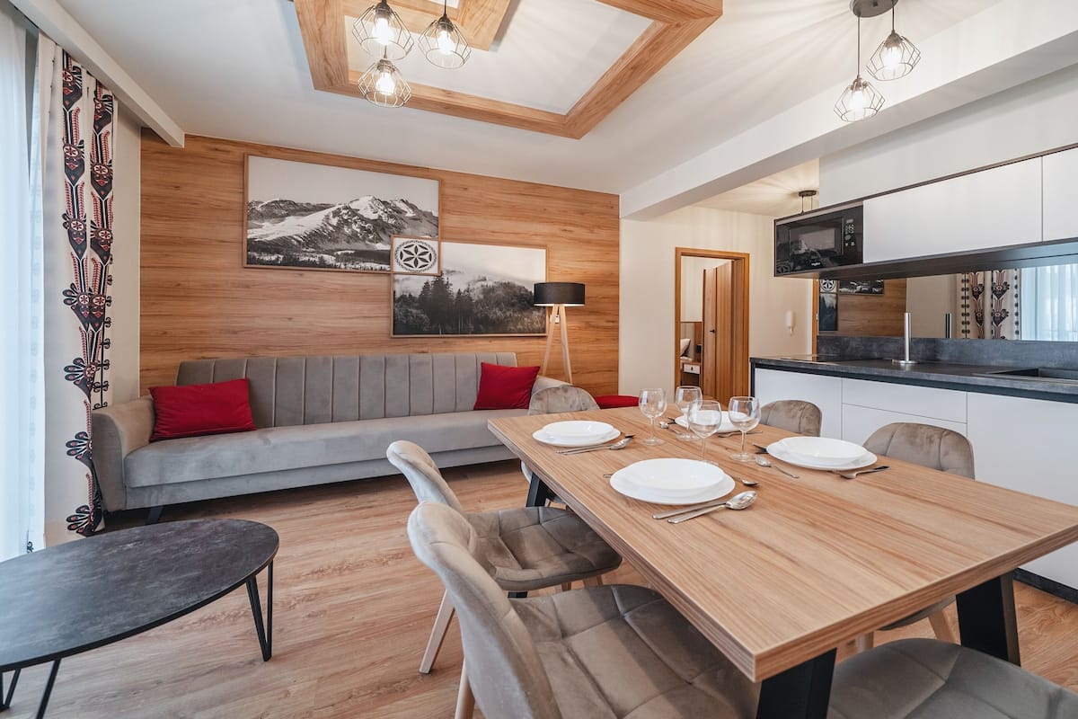 Ein modernes Wohnzimmer mit einem Holzespeisetisch, gedeckt für vier Personen, einem grauen Sofa mit roten Kissen und einem großen Fenster mit Vorhängen.