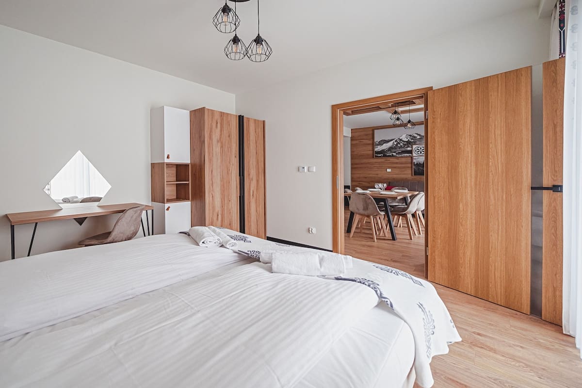 Ein modernes Schlafzimmer mit einem großen Bett, einem Schreibtisch und einem Essbereich, der durch eine offene Tür sichtbar ist.