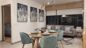 A modern dining area with a round table, light blue chairs, and a chandelier. There are abstract paintings on the wall, a sofa, and a view of the city at night through a large window.