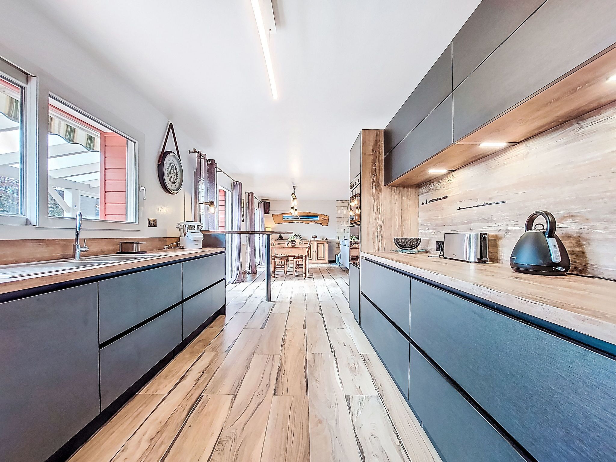 A modern kitchen with a wooden floor, dark cabinets, and a view into the dining area.
