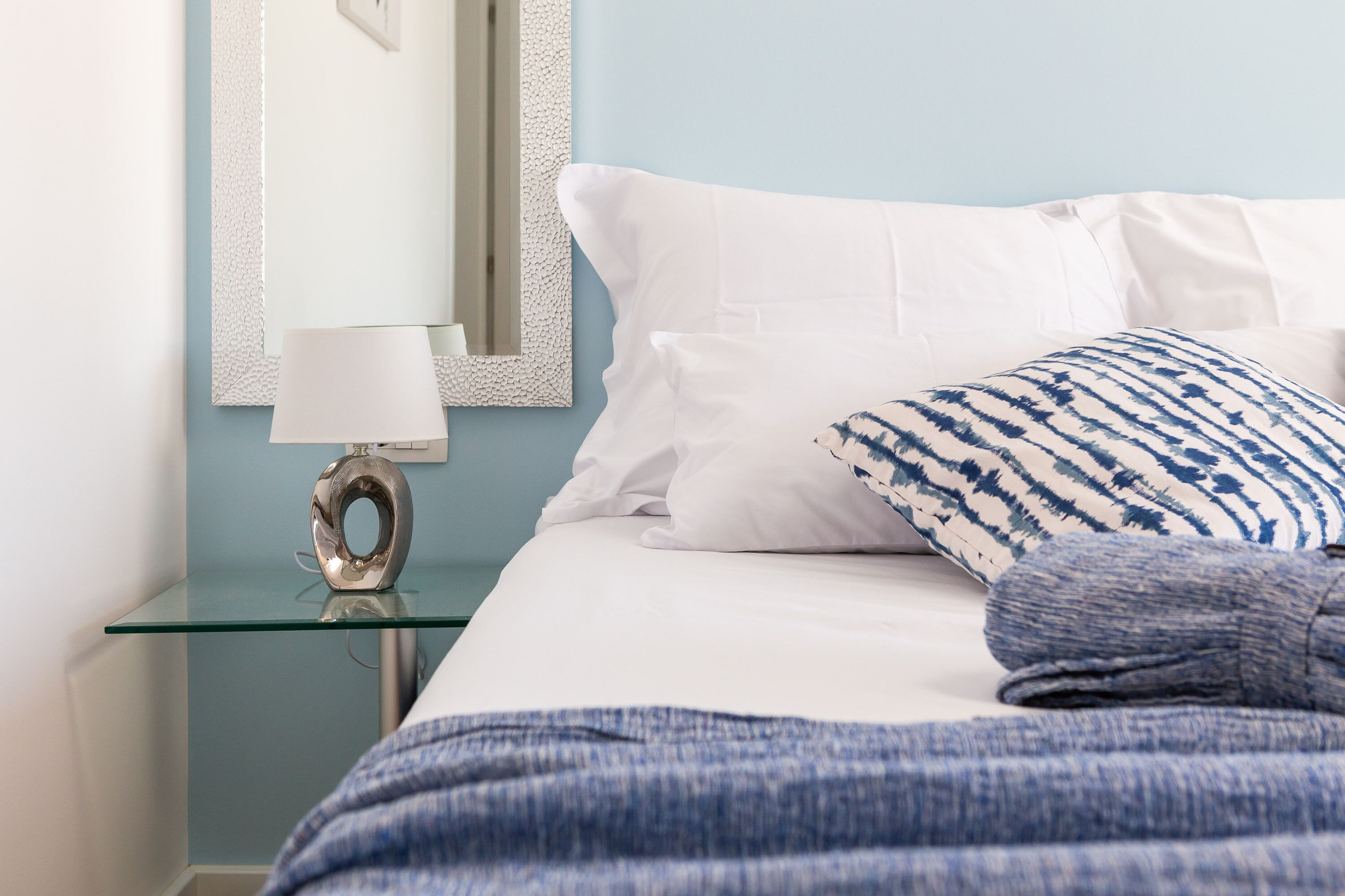 Una cama bien hecha con almohadas de diseño blanco y azul, una mesita de noche de vidrio con una lámpara moderna y una pared azul claro con un espejo enmarcado.
