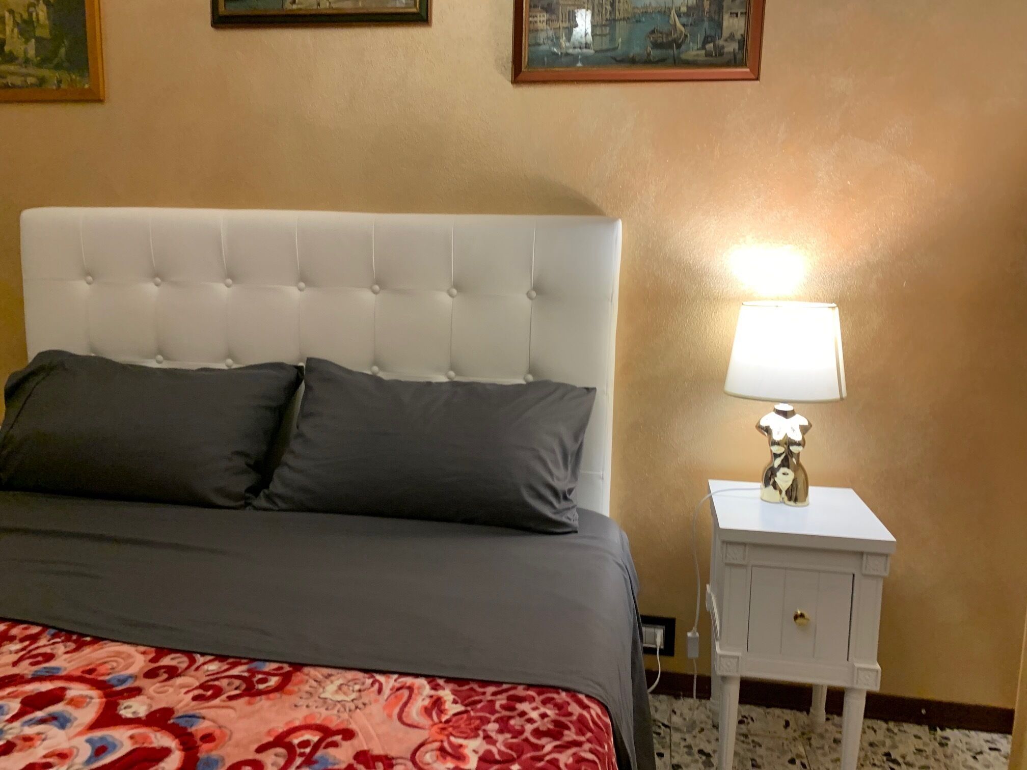 Ein Bett mit geblattetem Kopfteil, zwei Kissen, einer gemusterten Tagesdecke, einem Nachttisch mit Lampe und einer Wand mit zwei gerahmten Bildern.