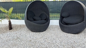 Deux chaises en osier noir avec des coussins, posées sur un sol de gravier, un petit palmier et une clôture verte en arrière-plan.
