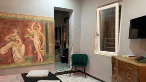 Une chambre d’hôtel avec un lit, un fauteuil vert, une commode en bois, un téléviseur et un grand tableau classique accroché au mur.
