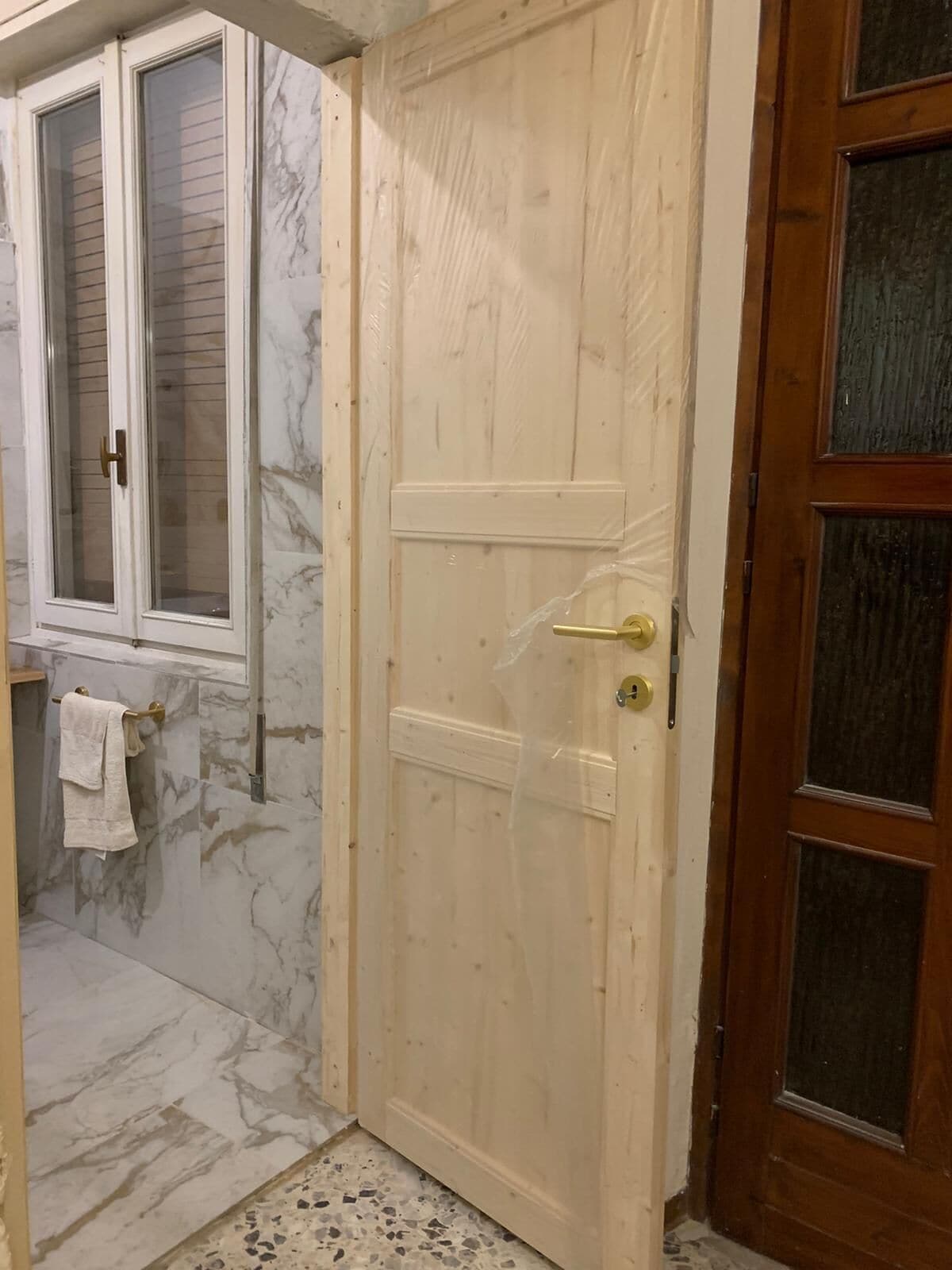 Eine Holztür mit Messingklinke, ein Fenster mit Jalousien und ein Boden mit Marmorfliesen.