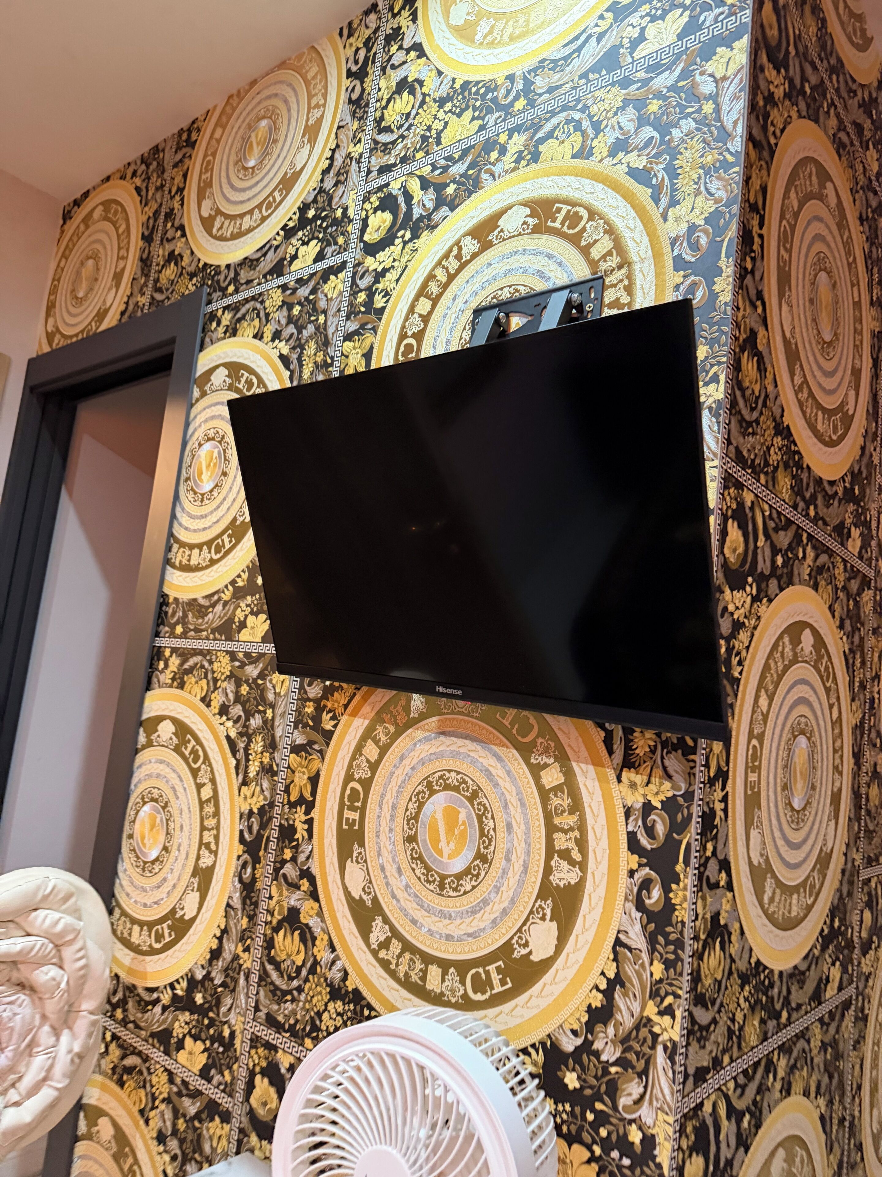 A flat-screen TV mounted on a wall with ornate wallpaper featuring circular patterns and the word 'GRACE.'