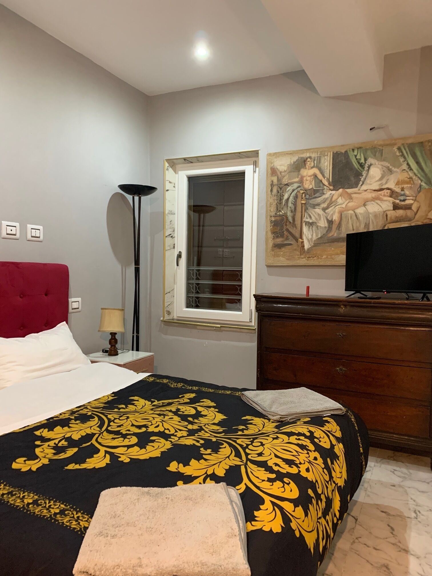 Una habitación de hotel con cama, cabecera roja, un cuadro, cómoda y televisor.