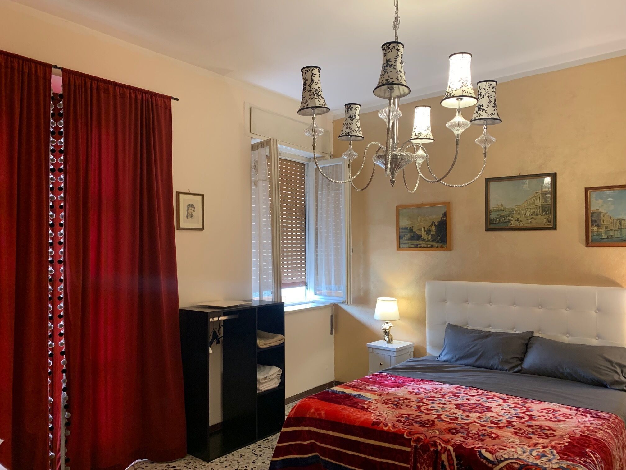 A bedroom with a bed, a chandelier, a window with blinds, and artwork on the wall.