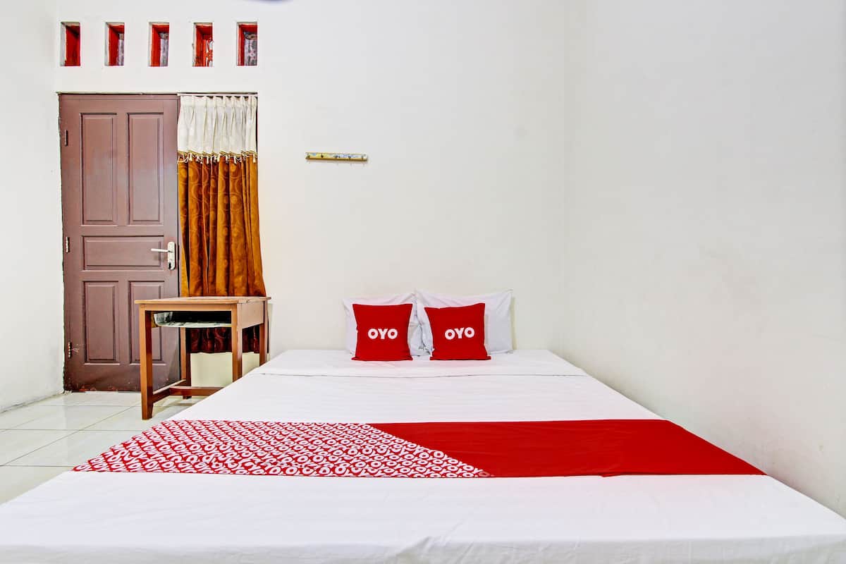 Een hotelkamer met een bed, twee rode kussens, een houten nachtkastje, een deken met een rood-wit patroon en een bruine deur.