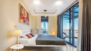 A modern hotel room with a large bed, a side table, a lamp, a painting on the wall, and a view of a building through large windows. - HB apartments Besik (Tbilisi)
