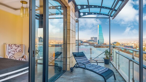 A balcony with a glass door, a lounge chair, and a view of a cityscape including a tall building. - HB apartments Besik (Tbilisi)