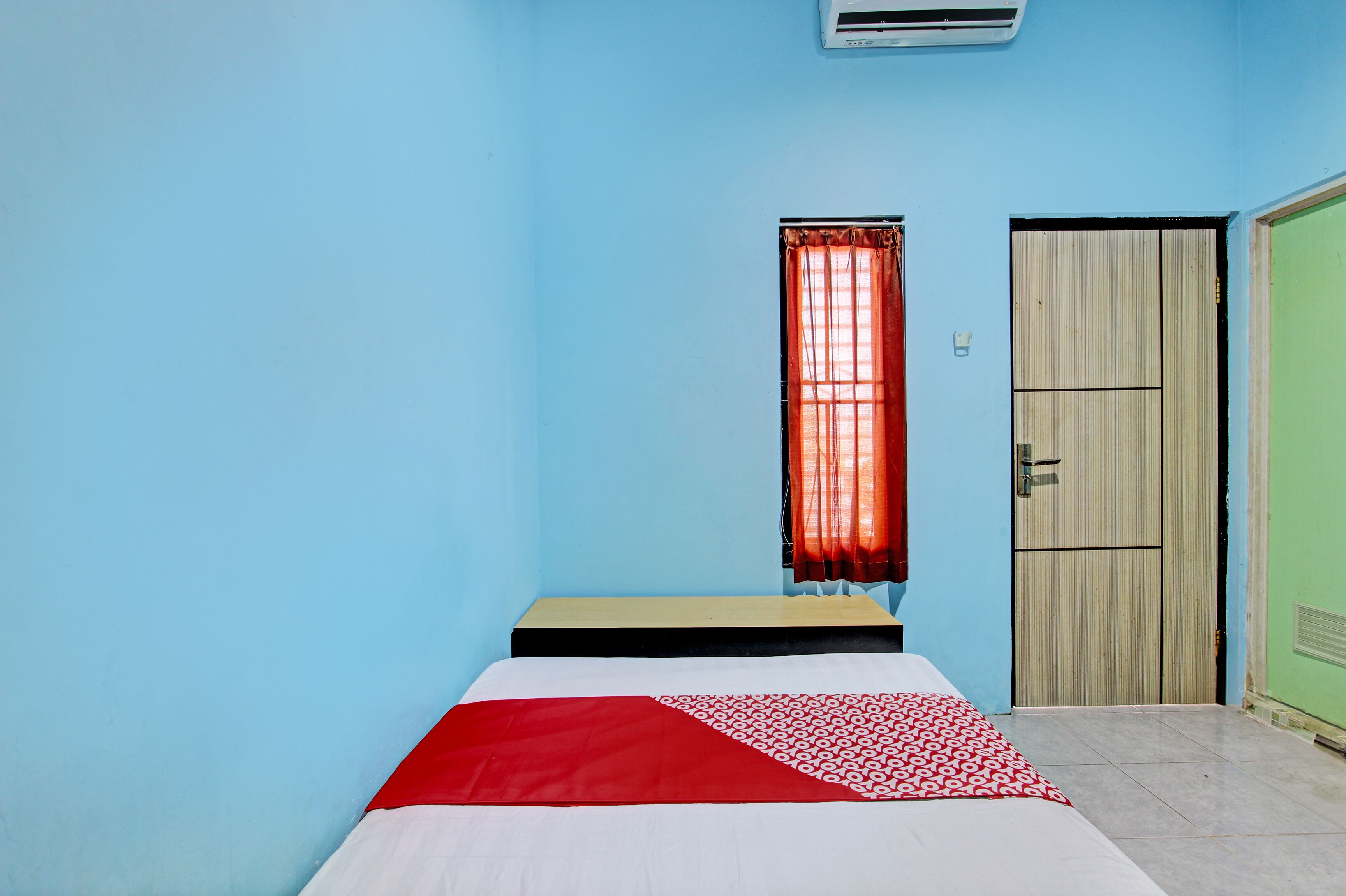 A room with a bed, a window with red curtains, a door, and an air conditioning unit.