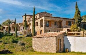 Traditional stone building with a rustic entrance and adjacent structure, set amidst a serene landscape with neatly trimmed hedges and tall trees.