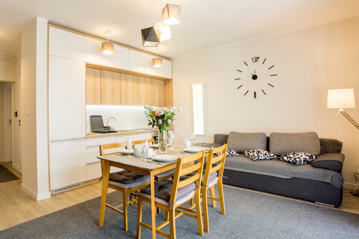Una cocina moderna con armarios de madera, una mesa de comedor para cuatro personas y una sala de estar con un sofá y un reloj en la pared.