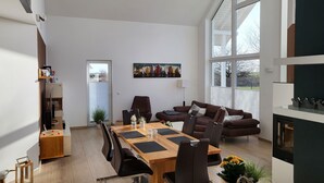A modern living room with a large dining table, comfortable seating, a fireplace, and a large window offering a view of the outdoors. - Modernes Ferienhaus mit Kamin, Garten und Eigener Sauna - Ferienhaus Meerfreude (Vaschvitz)