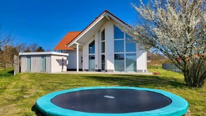 A modern house with a large glass window, a red roof, and a trampoline in the yard. - Modernes Ferienhaus mit Kamin, Garten und Eigener Sauna - Ferienhaus Meerfreude (Vaschvitz)
