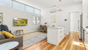 A modern kitchen with white cabinetry, a dining area with a gray sofa and a small round table, and a large window with blinds.