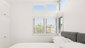 A bedroom with a bed, white walls, a window with shutters, and a grey headboard.