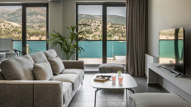 A modern living room with a sofa, a coffee table, and a TV. It features large windows with a view of a mountainous landscape and a body of water.