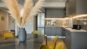 A modern kitchen with grey cabinets, a stainless steel refrigerator, and a dining area with a glass table and yellow chairs.