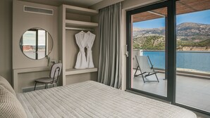 A modern hotel room with a large bed, a round mirror, a chair, and a view of a body of water and mountains. - Grand Terrace Suite/105m² - Spacious outdoor living area (Argostoli)