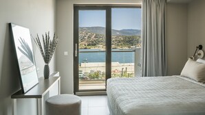 A bedroom with a bed, a desk with a framed picture, a vase with dried plants, and a view of the sea and hills through a large window. - Boutique Suite-2 bedroom-Sea View/100m² (Argostoli)