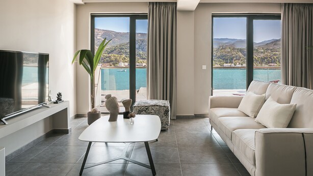 A modern living room with a large flat-screen TV, a white coffee table, a beige sofa, and a view of a mountainous landscape through large windows. - Luxury Living Suite-2 bedroom-Sea View/115m² (Argostoli)