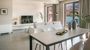 A modern living room with a sofa, dining table, and a TV. There are plants and a view of a mountainous landscape through large windows. - Luxury Living Suite-2 bedroom-Sea View/115m² (Argostoli)