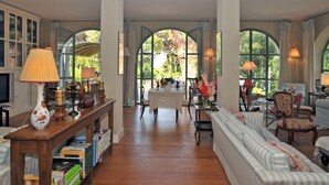 A spacious room with a wooden floor, a sofa, a dining table with chairs, and a large arched window providing a view of the outdoors. - Villa Gioconda - Homelike Villas (Bibbona)