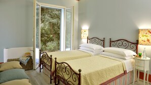 A bedroom with two beds, a window with a view of trees, a bedside table with a lamp, and a wicker basket. - Villa Allegra - Homelike Villas (Bibbona)